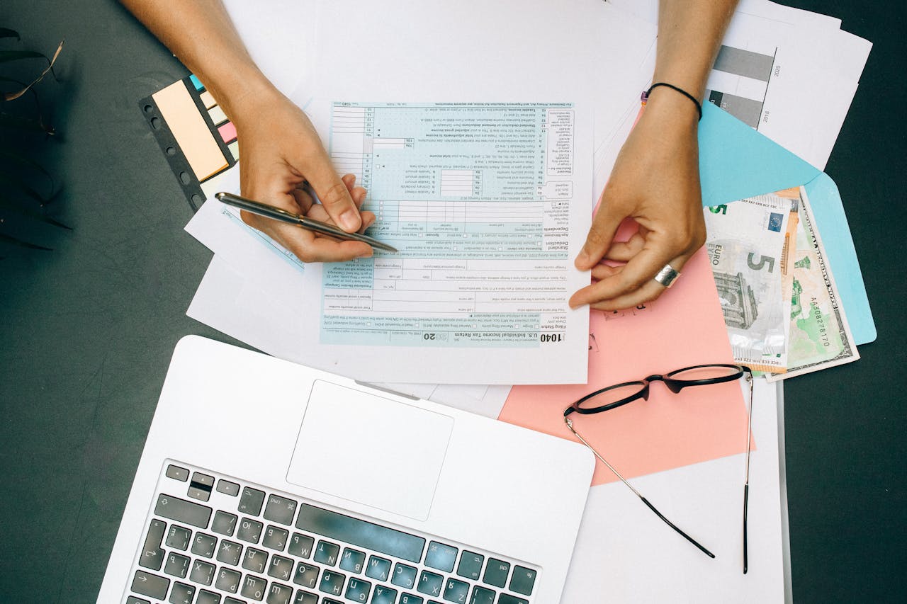 digital Hands writing on tax documents with laptop, glasses, and currency on desk.